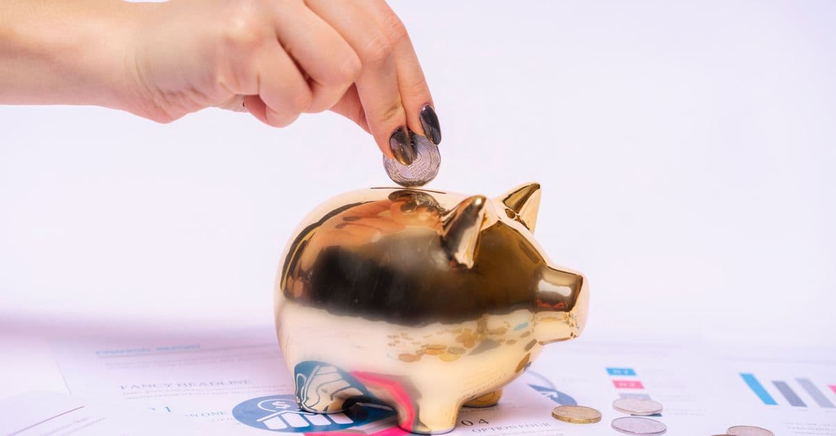 Eine Hand wirft eine Münze in ein goldenes Sparschwein auf Finanzunterlagen.