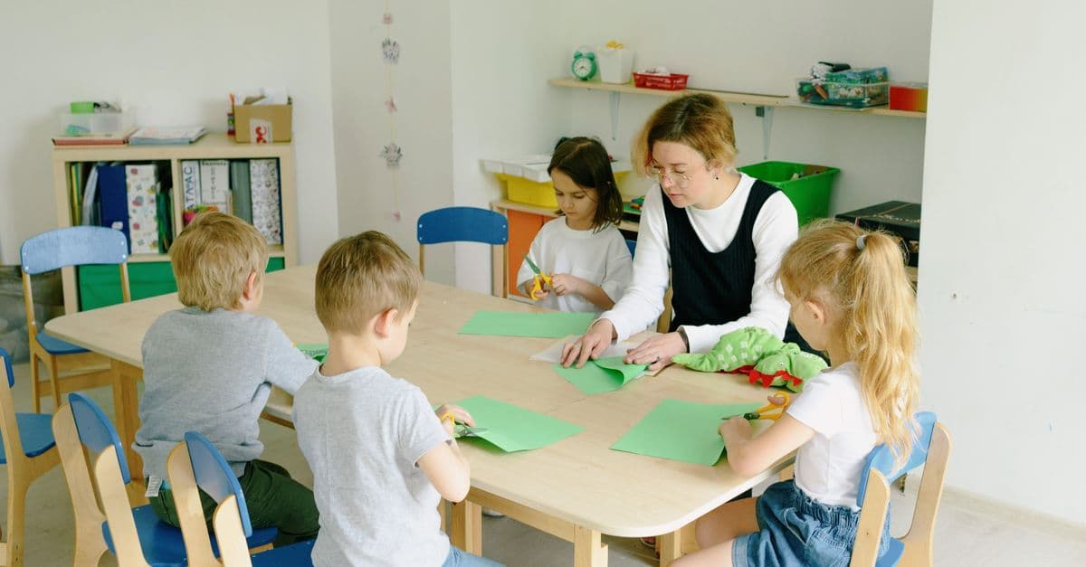 Kinder und Erzieherin basteln mit Papier und Scheren im Kindergartenraum.