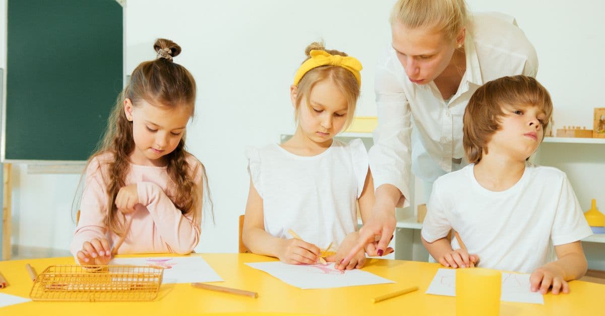 Lehrkraft unterstützt junge Kinder beim Zeichnen in einem hellen Klassenraum.