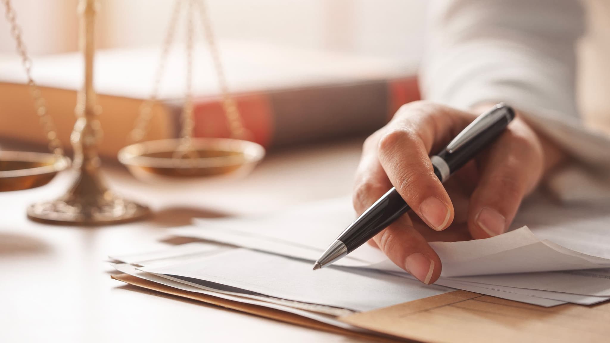 Eine Person schreibt an einem Schreibtisch auf juristischen Dokumenten, im Hintergrund unscharfe Justizwaage.