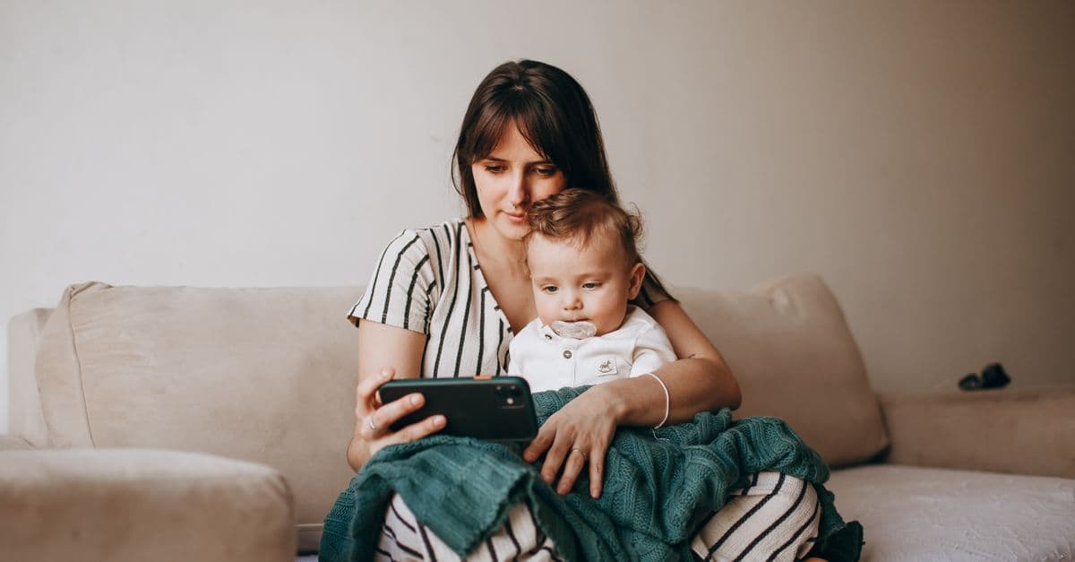 Mutter und Baby sitzen auf einem Sofa und schauen gemeinsam auf ein Smartphone.