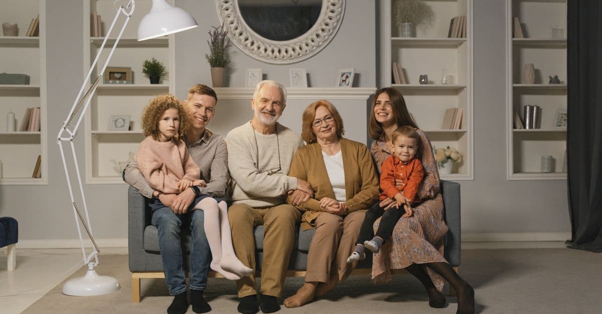 Mehrgenerationenfamilie sitzt gemeinsam auf einem Sofa in einem warmen Wohnzimmer.