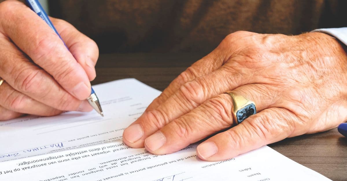Nahaufnahme einer älteren Person beim Unterschreiben eines Dokuments, mit Fokus auf Hand und Goldring.