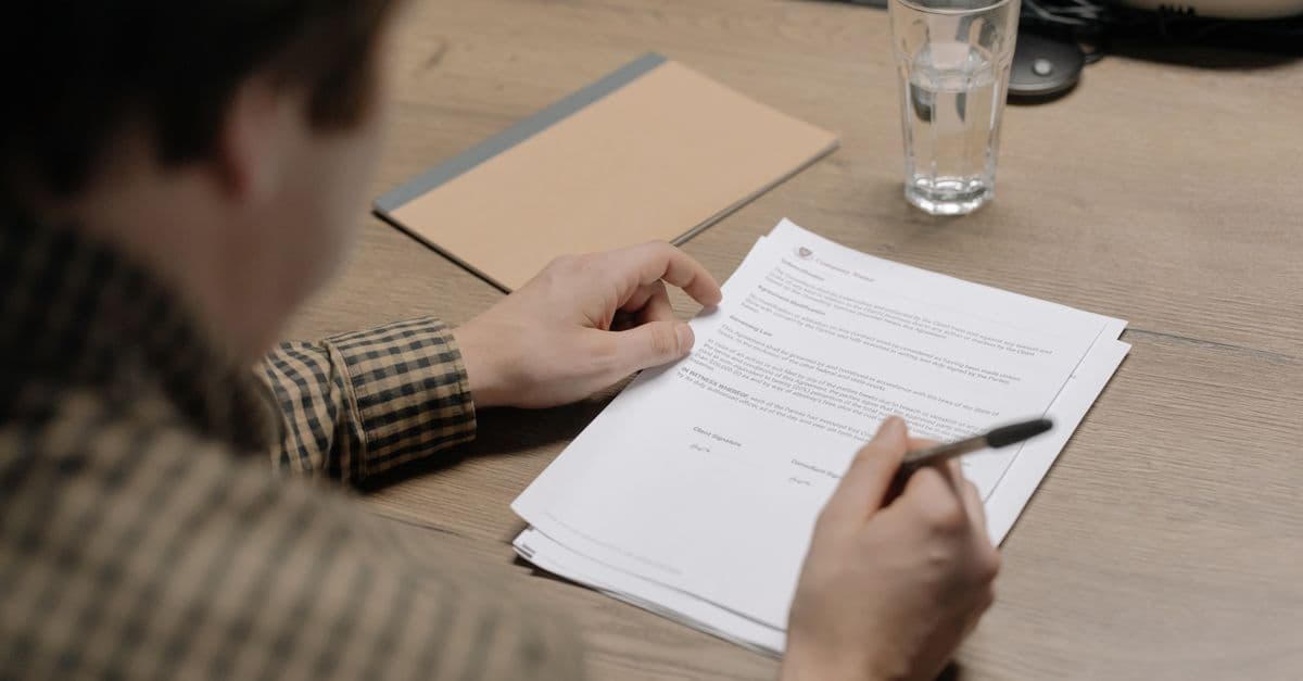 Geschäftsmann prüft einen Vertrag am Schreibtisch und hält einen Stift, daneben steht ein Glas Wasser.
